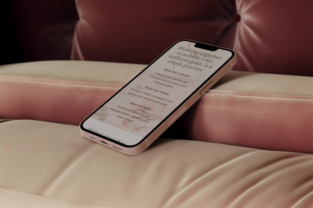 A smartphone with a wellness app displayed on the screen rests on a beige couch. The background features a plush, rose-colored cushion. The app includes text about achieving wellness goals and options for booking services with a massage therapist, all wrapped in an elegant showit template design.