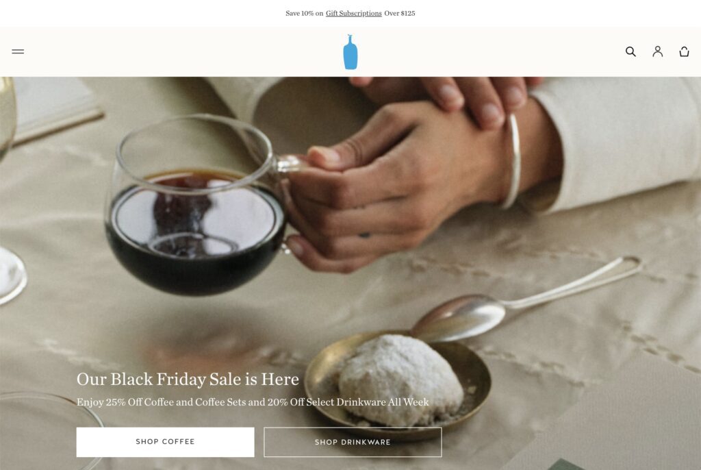 A website homepage featuring a close-up of a hand holding a glass mug of black coffee, placed on an elegant table setting with a plate of powdered cookies and a silver spoon. 