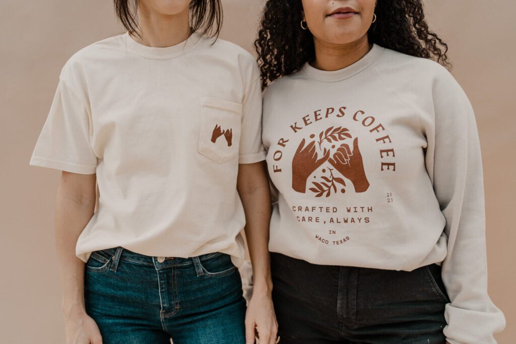 For Keeps Coffee-branded apparel featuring a beige t-shirt with a pocket design and a beige sweatshirt with the full logo. Both designs use earthy brown tones and emphasize the shop's “Crafted with Care, Always” tagline.