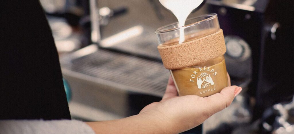 A creamy latte in a branded cup for For Keeps Coffee, featuring the logo with hands and coffee branches. The cup is placed on a marble surface alongside smaller cups of coffee in the background.