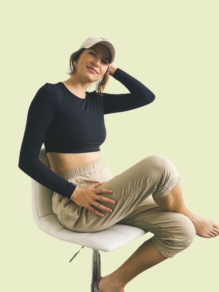 Caitlin Christensen, founder of Creative SEO Coach, sitting on a white chair, wearing a casual black crop top, beige pants, and a light-colored cap, smiling confidently against a light green background.